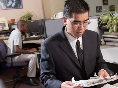 A young man in a suit works at a desk, while a co-worker files things behind him