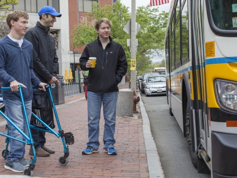Three men wait on the sidewalk as a bus pulls up. One of them is using a walker.