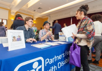Two men sitting behind a table that has flyers on it, talking to a college-aged woman standing by the table.