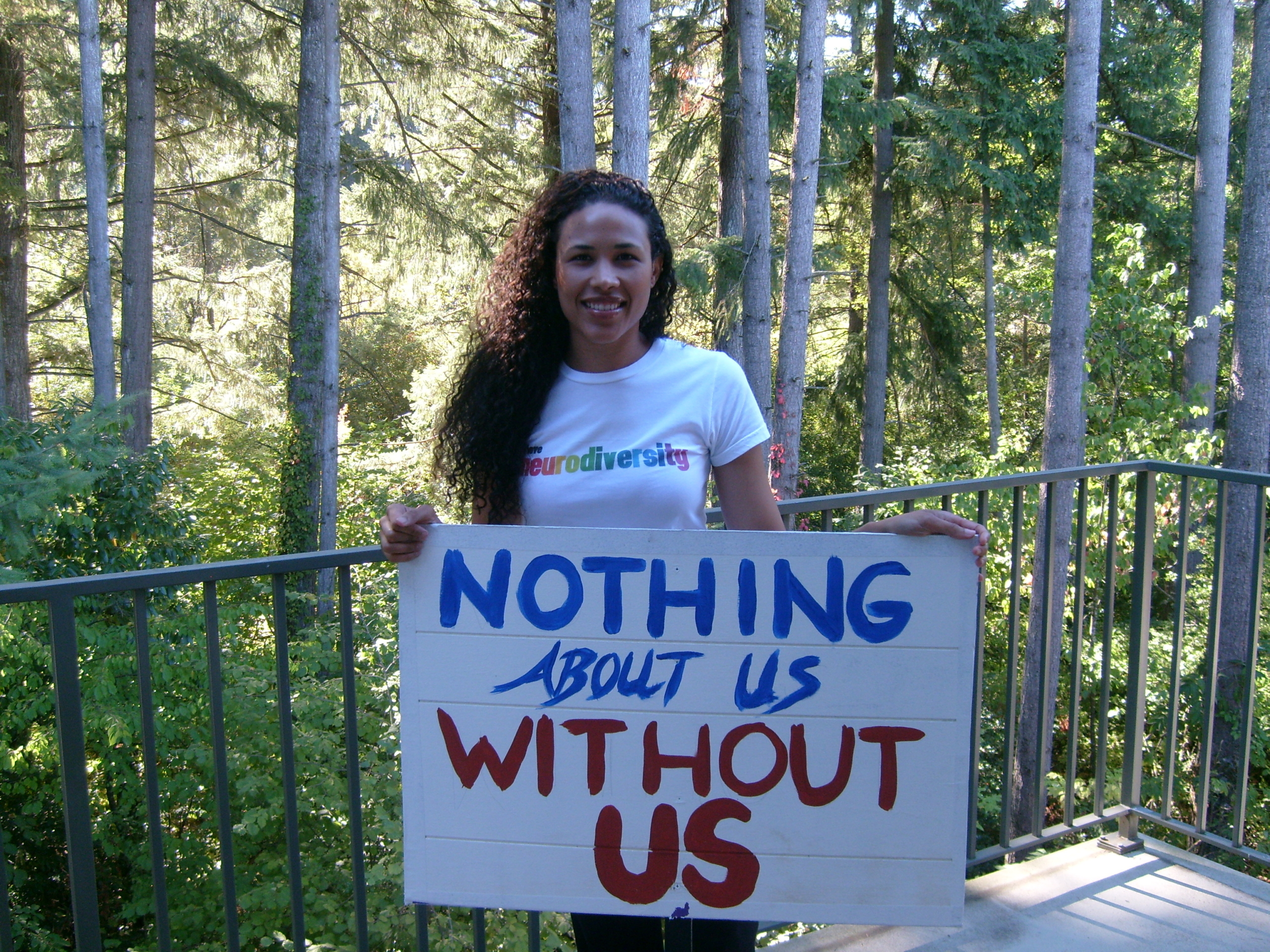 Young woman wearing a shirt that says Neurodiversity and holding a sign saying Nothing About Us Without Us