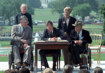 George H.W. Bush signs the ADA into law, with two disabled advocates and lawmakers seated next to him in their wheelchairs