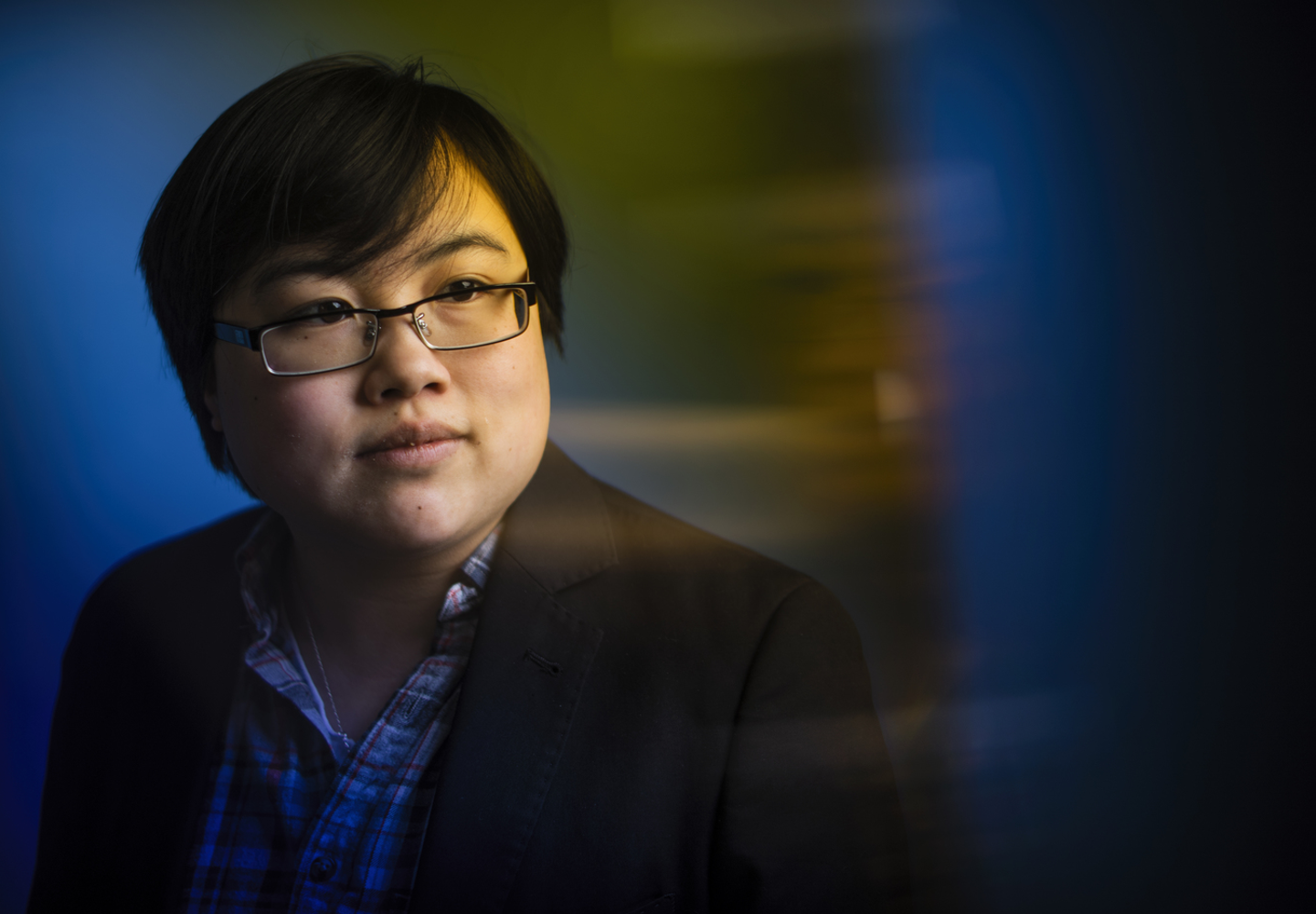 Photo: Headshot of Lydia Brown, young East Asian person, with short black hair, wearing glasses, a plaid shirt, and black jacket. They are looking in the distance, posed against a stylized blue dramatic background. Photo by Adam Glanzman.