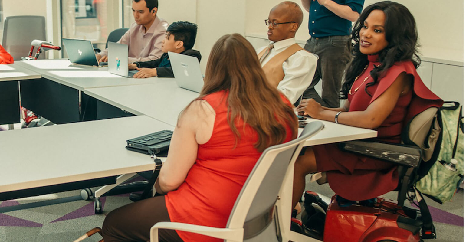 An image of people meeting in a conference room. The tables in the room are set up in the shape of the letter U. The person in the center of the image is a white person, they have an accessible keyboard sitting on the table, and a black service dog lying at their feet. The person to their right is a black person using a motorized wheelchair, the two people seem to be speaking to one another. There are three other people in the background doing work on their laptops. There is one more person standing behind the group.