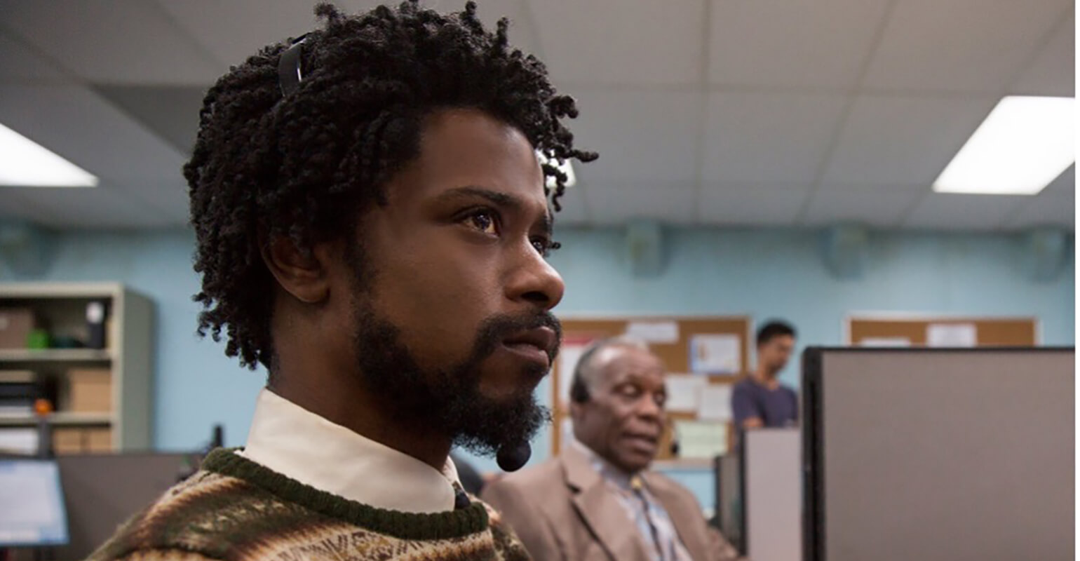 An image of actor LaKeith Stanfield in the movie “Sorry to Bother You.” He is sitting in a call center looking into the distance. Behind him are coworkers that appear to be taking calls for their employer. The movie is an absurdist comedy that heavily features a theme of code-switching.
