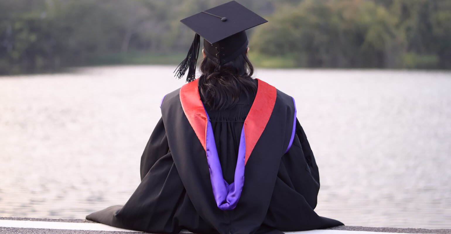 An image of a person in a graduation cap and gown sitting on a ledge. They are looking over a lake with a line of trees on the other side of the water.