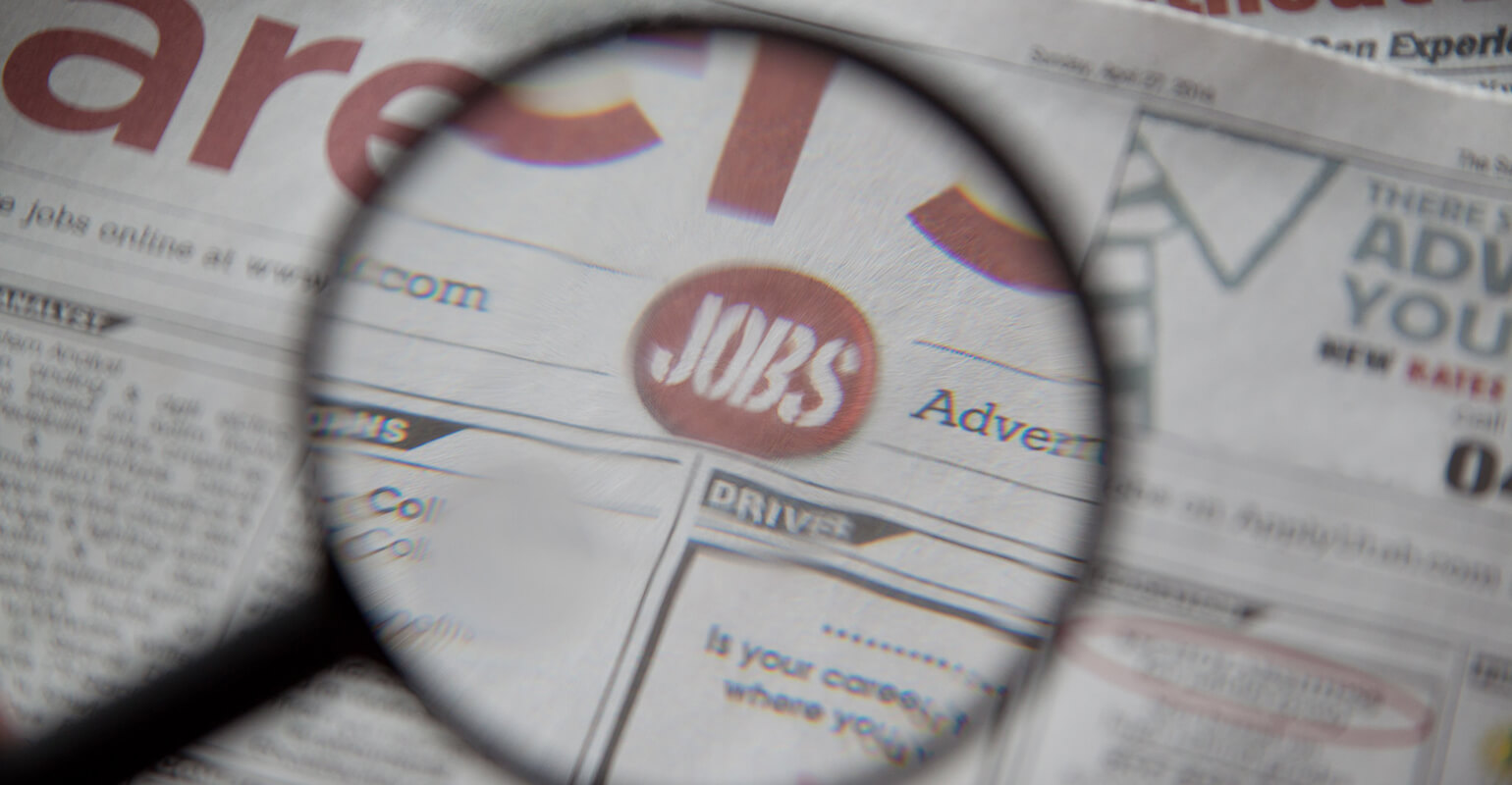 An image of a magnifying glass focusing on a newspaper. The magnifying glass is focused on the word jobs.