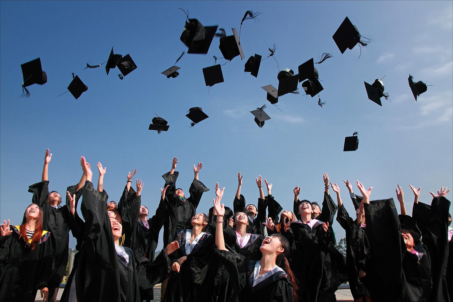 A large group of students wearing black graduation gowns. They are outside, throwing their caps into the air, celebrating their graduation.