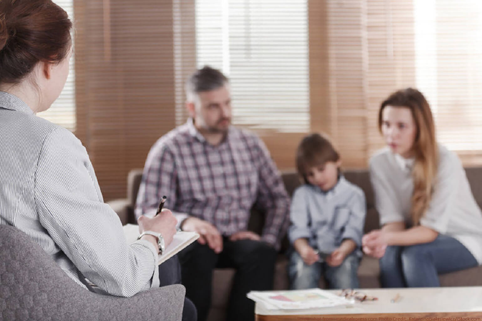An image of a family therapy session. On the left, a therapist sits in a chair taking notes on a clipboard. On the opposite end of the room, out of focus, a family sits with a small child in the middle. In front of the family, there is a table with some of the child’s drawings