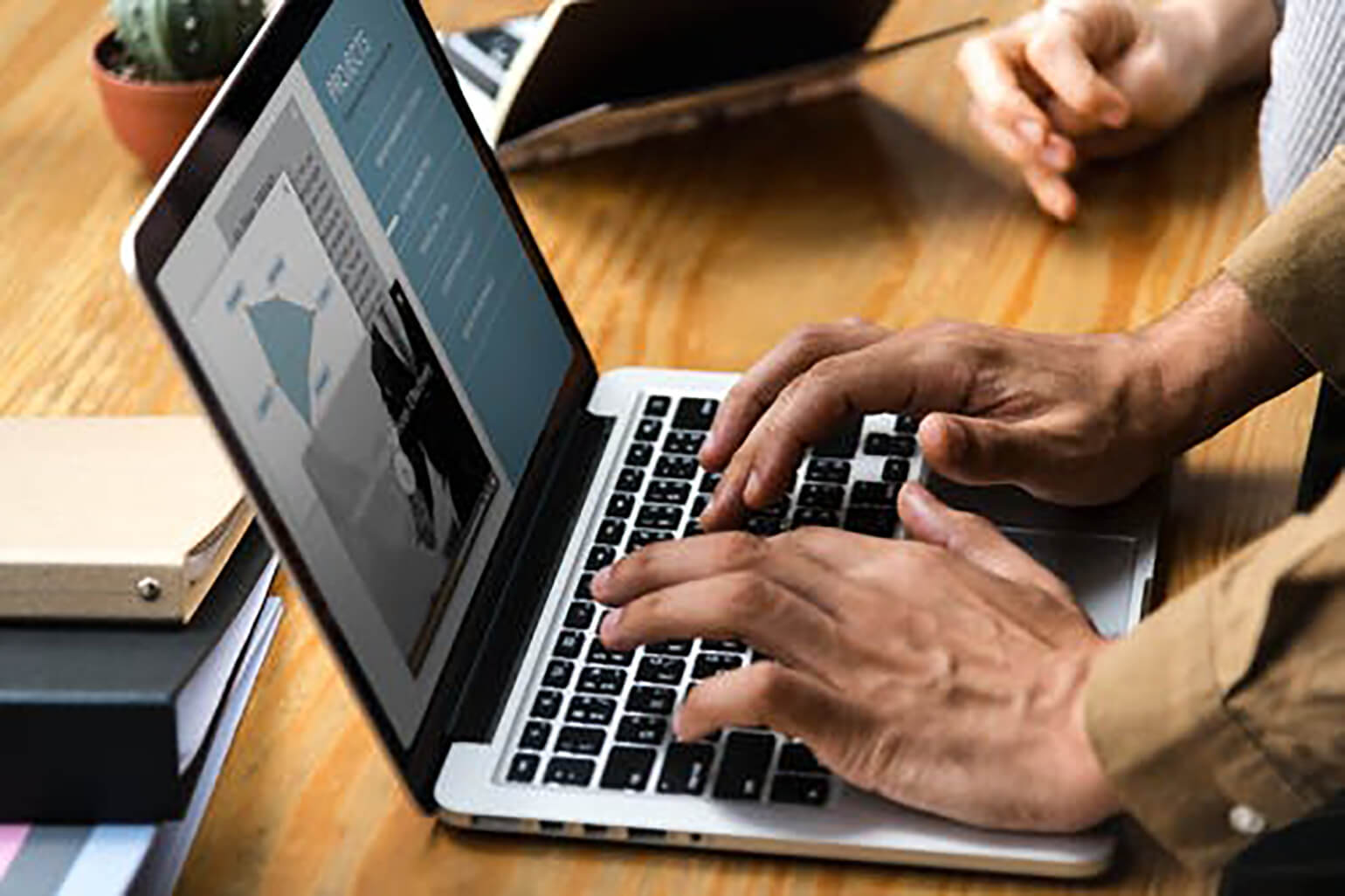 Image of hands typing on an open laptop that is on a wooden table or desk. Nearby are another set of hands leaning on the desk.