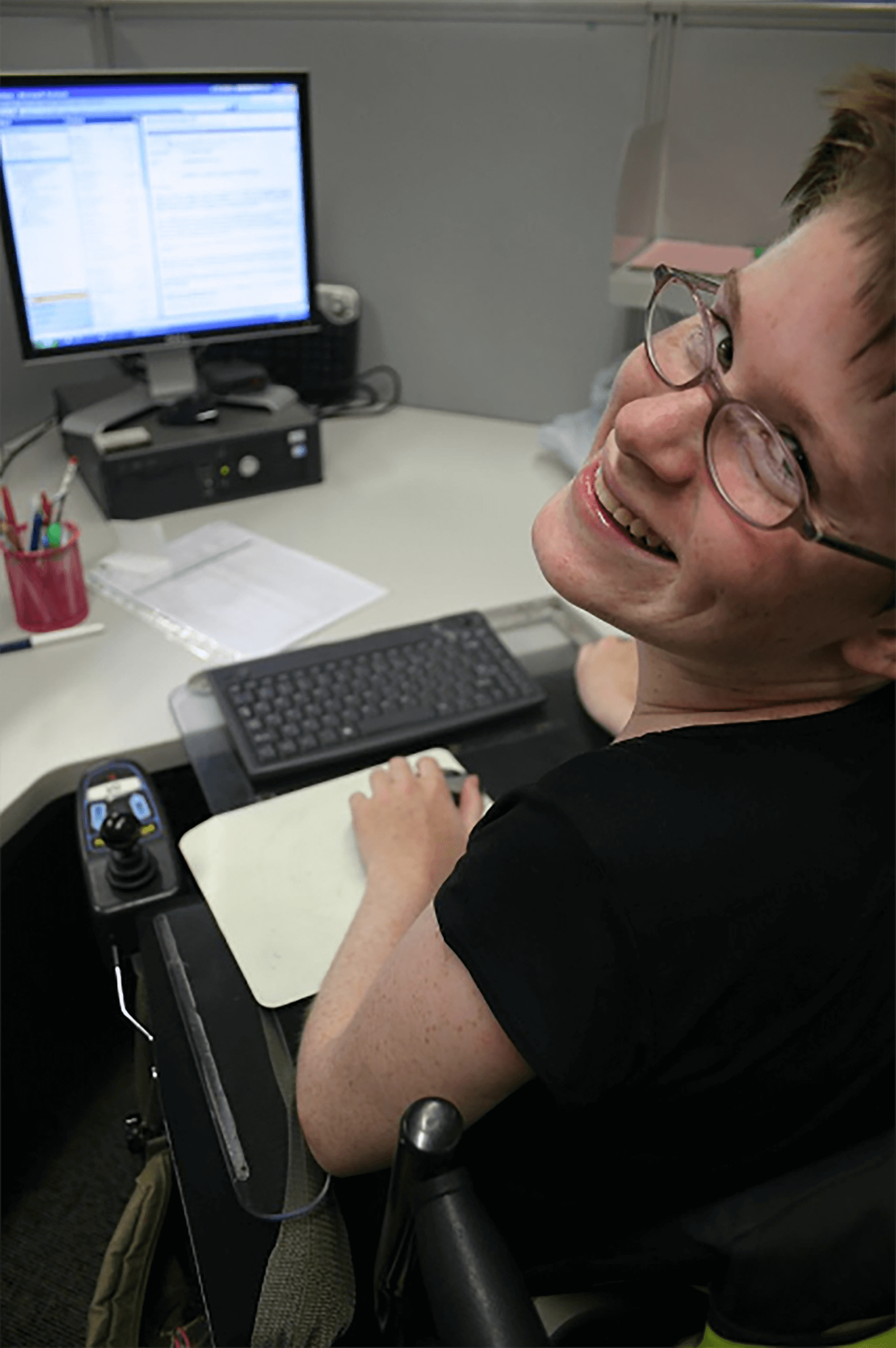 A person with glasses and short, brown hair looks over their shoulder into the camera and smiles. They are working at a desk with a computer on it. They are sitting in a wheelchair with a keyboard on a lap desk in front of them.
