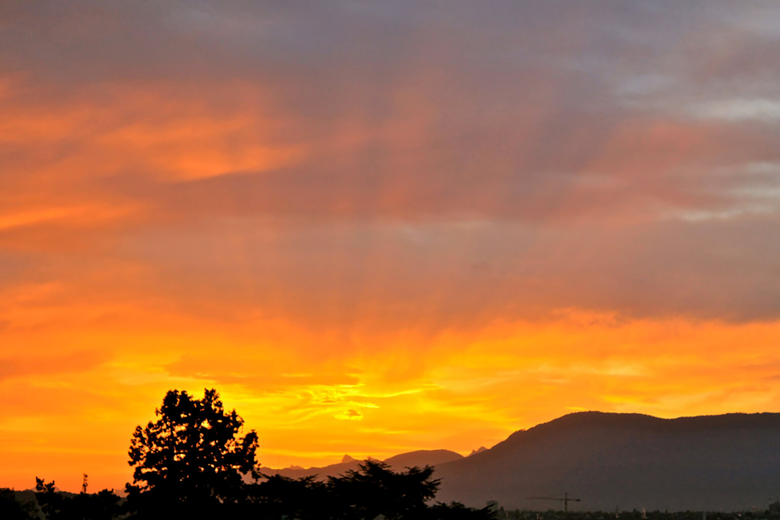 This is a photograph of an orange and pink and gray sunrise in the sky, over some mountains and treetops that are still in shadow.