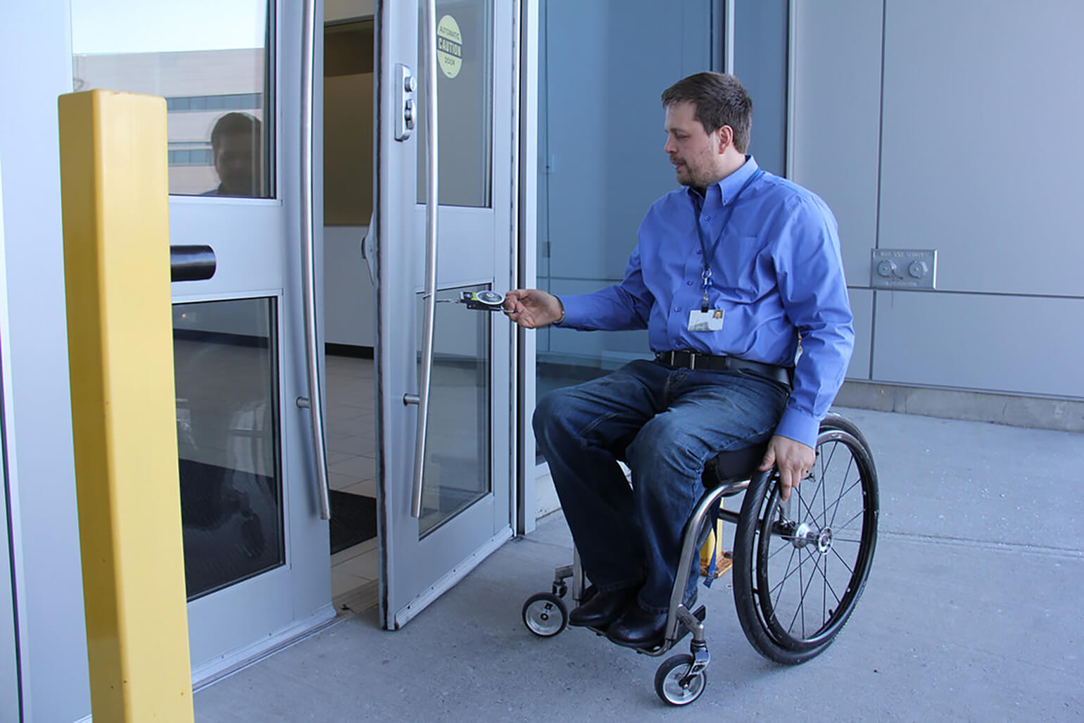 An image of a person using a wheelchair opening a door. They are using a handheld hook to pull a door open, rather than using a door handle or an accessibility button.