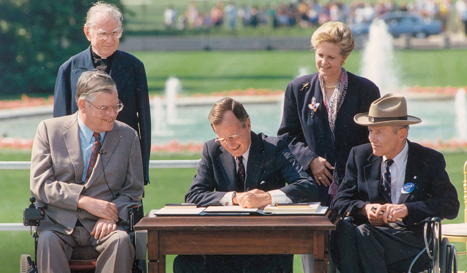 An Image of President George H.W. Bush signing the ADA into law in 1990. On his left, Evan Kemp sits in his wheelchair while Reverend Harold Wilke stands behind him. On his right, Justin Dart sits in his wheelchair and Swift Parrino stands behind the President.