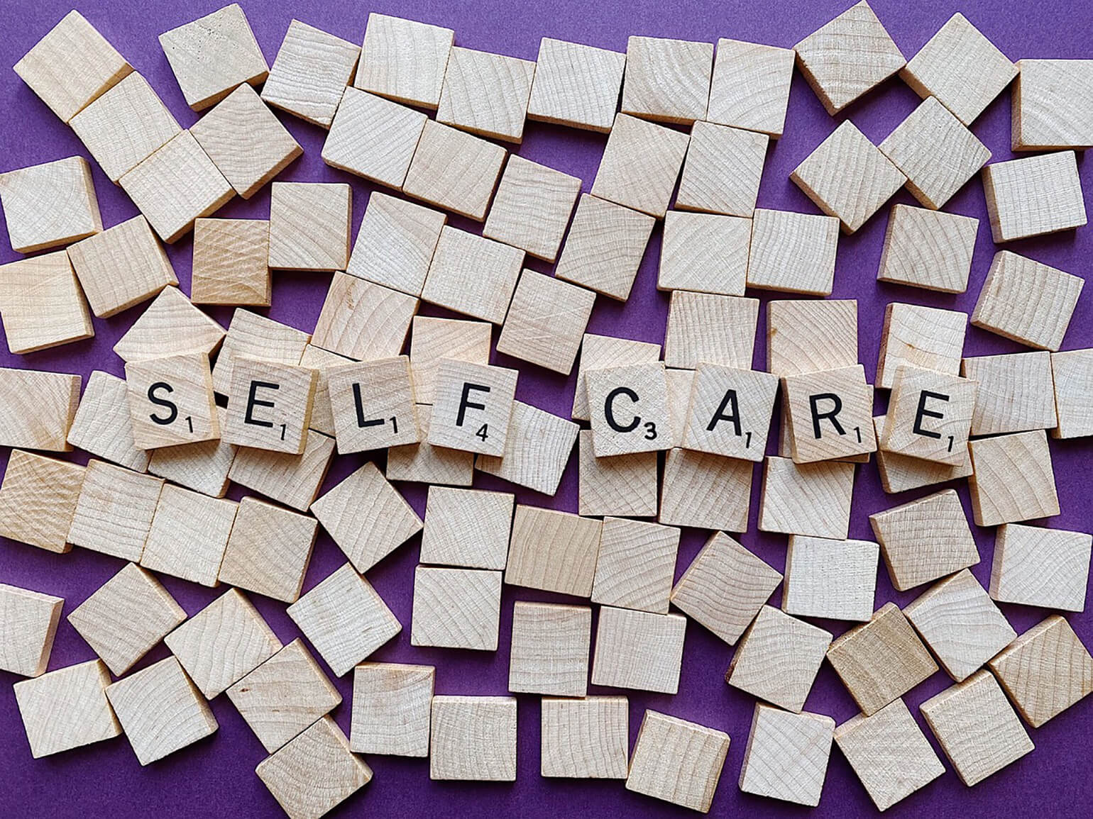 Image of wooden scrabble pieces scattered across a purple surface. Eight scrabble pieces are turned over in the middle of the pile to spell the words 'self care'.