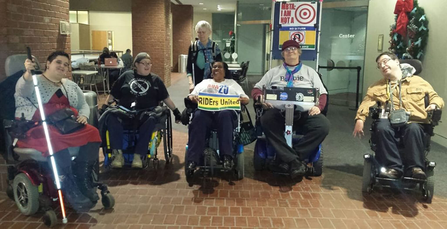 An image of 6 Members of the Boston Center for Independent Living protesting for increased accessibility on Boston public transportation. 5 of them sit in motorized wheelchairs, while 1 stands behind the group. The person in the middle holds a sign that says, 'RIDErs United.'